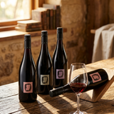 Three dark wine bottles with pink labels are arranged on a rustic wooden table beside a filled wine glass, with a third bottle lying on its side. The setting is indoors near a window with warm natural light and stone walls in the background.