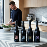 Four dark wine bottles with minimalist square labels in pink, gray, gold, and purple, arranged side by side on a white background.