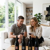 A man and a woman sit on a white sofa in a bright living room, each holding a glass of red wine while looking at four dark wine bottles lined up on a wooden coffee table in front of them.